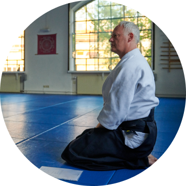 Ein Aikido Trainer mit einem weißen Trainingsanzug und schwarzer Hose kniet auf blauen Matten.