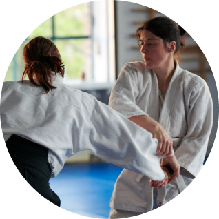 Zwei Frauen die Aikido machen. Eine der Frauen scheint die andere am Arm zu boden zu reißen.